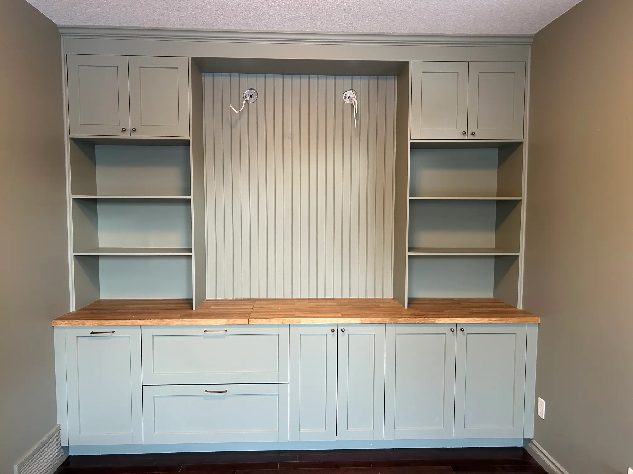 A built-in shelving unit with light green cabinets and wooden countertops. The central section features paneling with two hooks, and there are open shelves on either side. The room has light brown walls and dark flooring.