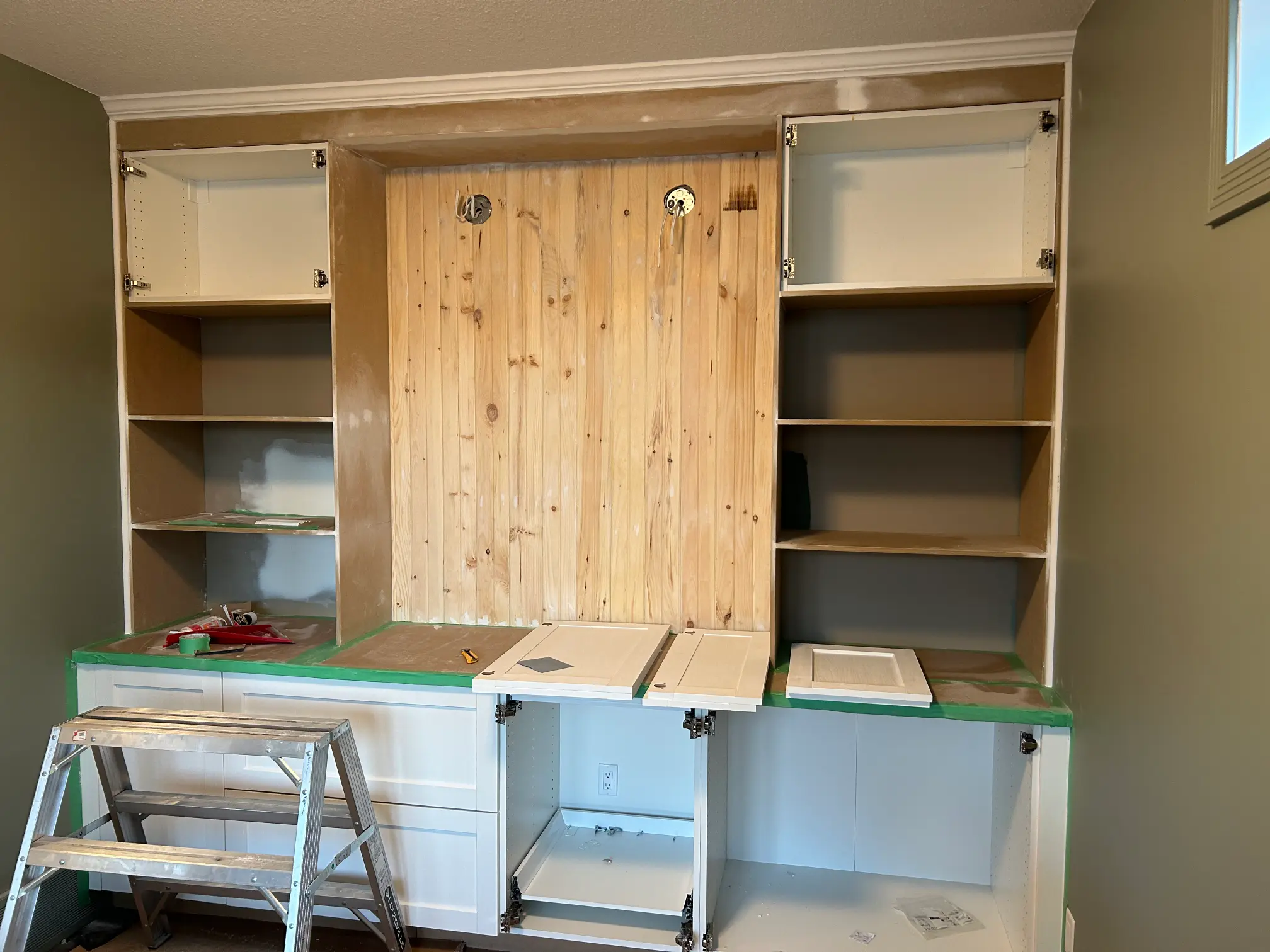 Partially assembled built-in cabinets in a room under renovation. The lower section has open doors, with tools and cabinet doors on top. Shelves on either side and a wooden back panel are visible. A ladder stands in front. Walls are painted beige.