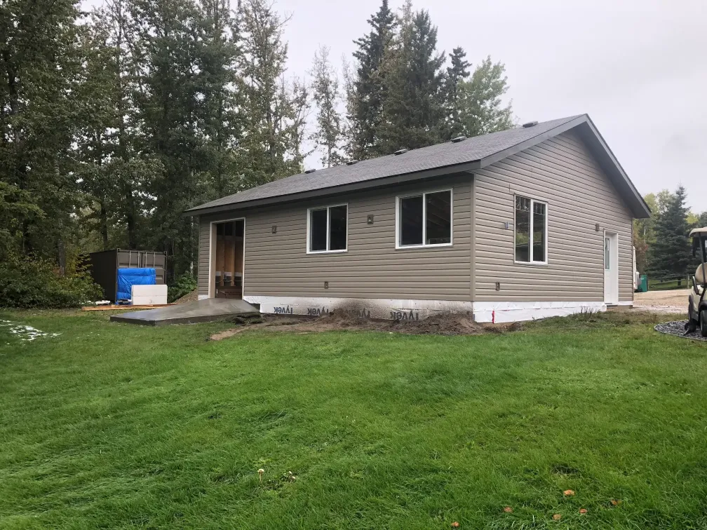 Small single-story house with beige siding, surrounded by a grassy lawn and tall trees. The roof is dark gray, and there is a cement walkway leading to the front door. The scene is overcast, suggesting a cloudy day.