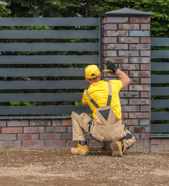 Worker in a yellow uniform installing a modern metal fence on a brick base.