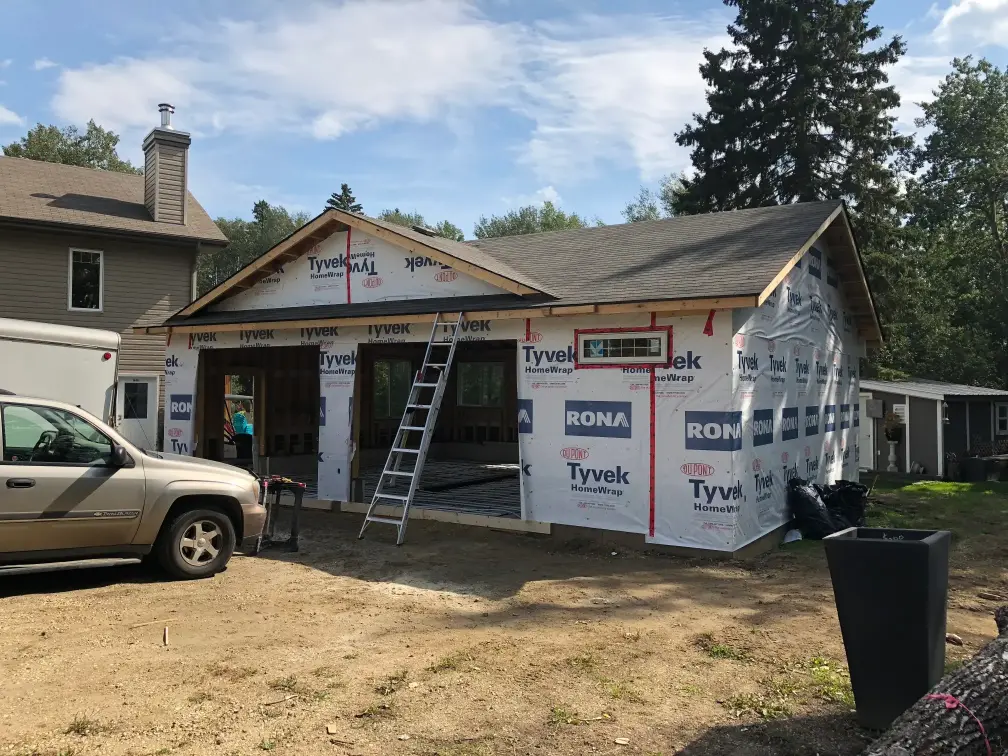 A partially constructed house with Tyvek insulation wrap is in the foreground. A ladder leans against the roof. A tan SUV is parked nearby. Trees and a neighboring house are visible in the background under a clear sky.