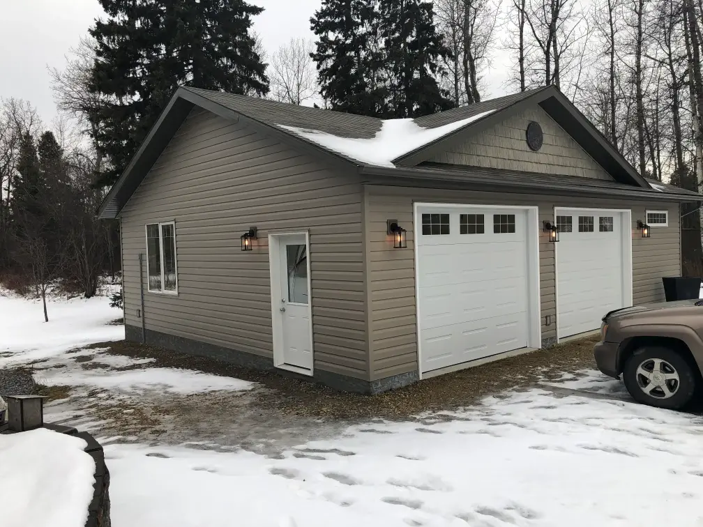 A beige detached garage with two white doors stands in a snowy landscape. It is surrounded by tall trees, and there is a vehicle partially visible on the right. The roof is covered in patches of snow.