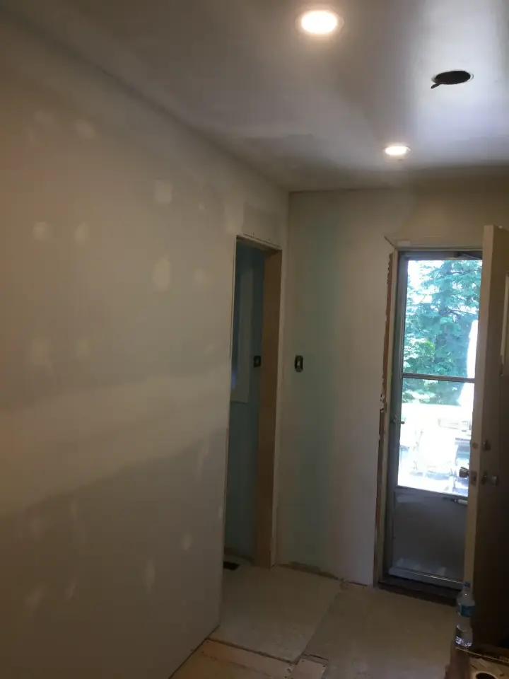 A partially completed interior hallway with drywall partially painted. Recessed lights are on the ceiling, and a door with a glass window leads outside, showing trees in the background. The floor is bare, and construction materials are visible.