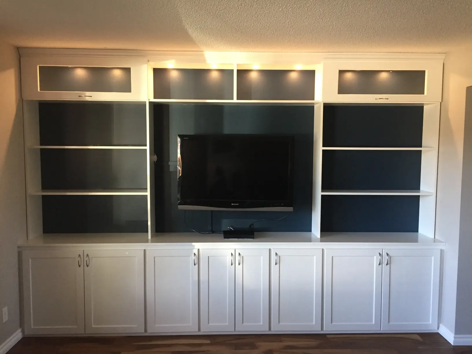 A white built-in entertainment center with illuminated shelves, featuring a mounted flat-screen TV in the center. The unit has cabinets below and open shelving on either side, set against a dark background. The floor is hardwood.