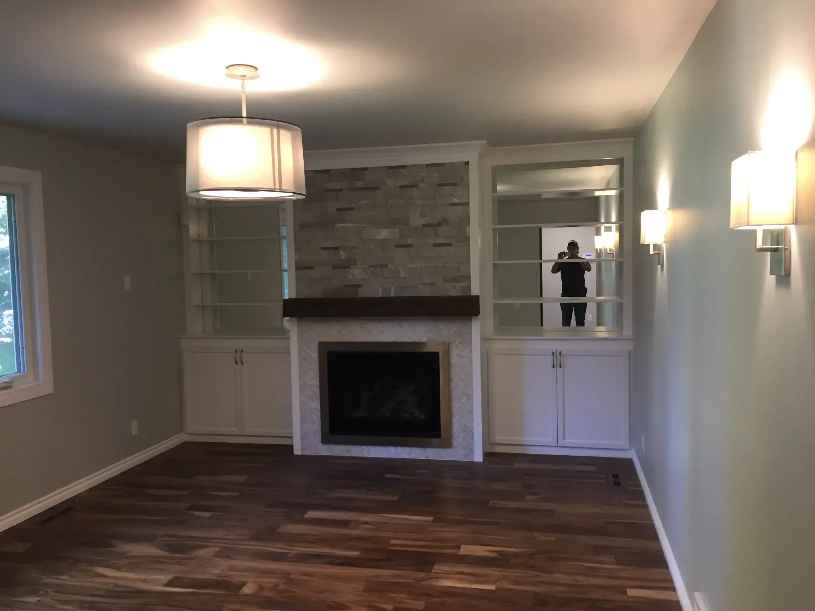Living room with wooden flooring, a modern chandelier, and a fireplace with stone detailing. Built-in shelves flank the fireplace. Wall sconces provide additional lighting. A large window lets in natural light.