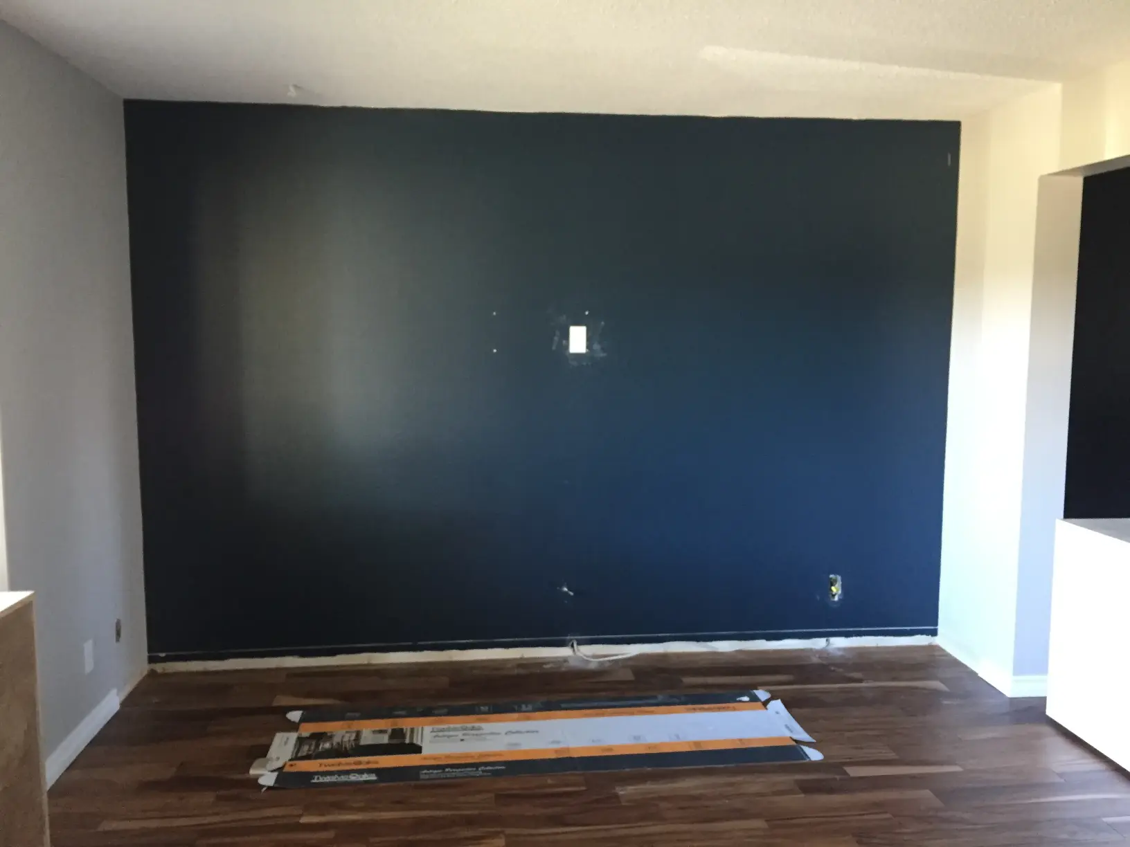 A room with a newly painted dark blue accent wall and wooden flooring. A protective sheet with paint supplies is laid out on the floor in front of the wall. The room is unfurnished and in progress of decoration.