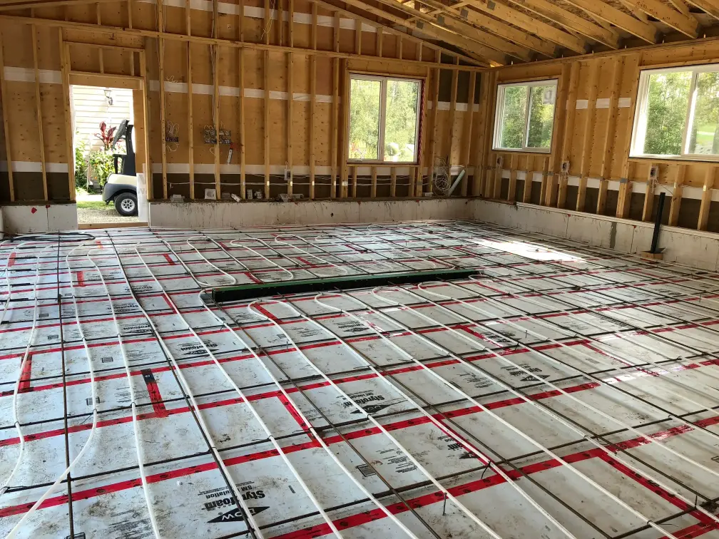 A room under construction with exposed wooden walls. The floor features a grid of white radiant heating tubes arranged in rows. There are several large windows, and sunlight is streaming in, illuminating the space.