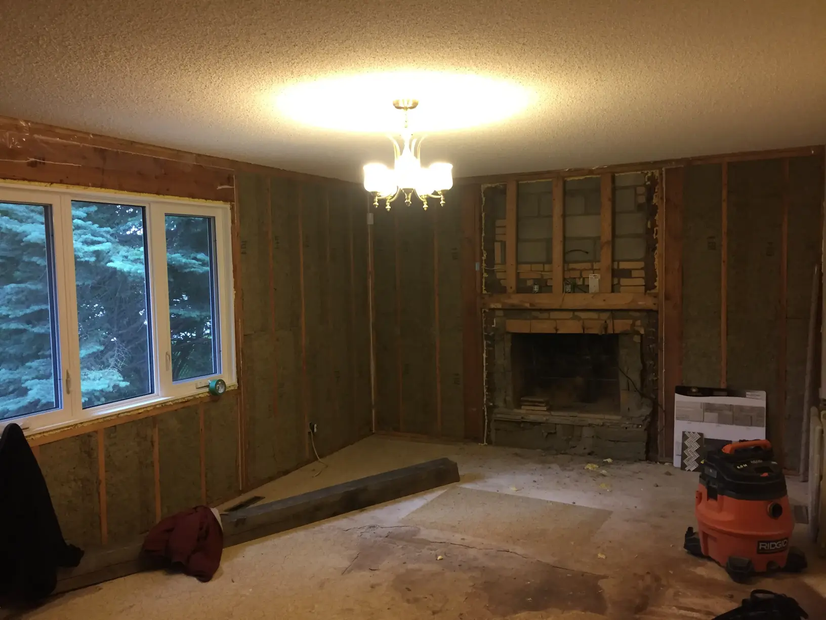 A dimly lit room under renovation with exposed wooden wall framing and insulation. A window shows trees outside. A bare light fixture hangs from the ceiling. A fireplace is partially dismantled, and a vacuum cleaner is on the carpeted floor.