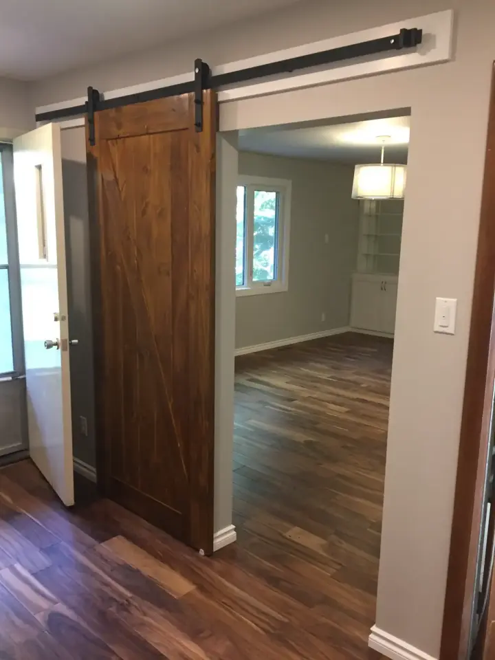 A room with wooden flooring and a sliding barn door. The doorway leads to another room with large windows and a ceiling light fixture. Walls are painted light gray, and there is a white light switch on the right.