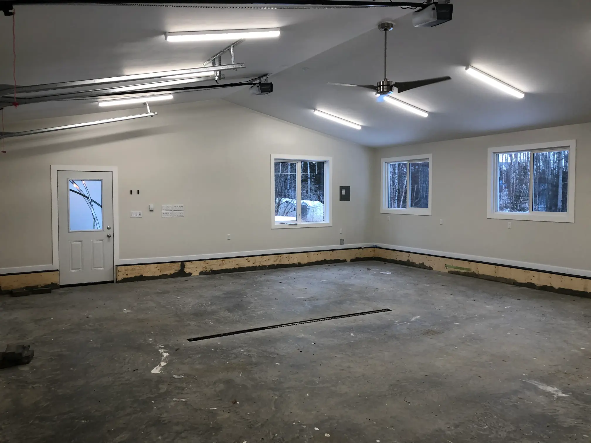 A large, empty garage with clean concrete floors and white walls. It features fluorescent ceiling lights, a ceiling fan, three windows, and a white door. The room is spacious and undecorated, with a snowy outdoor scene visible through the windows.